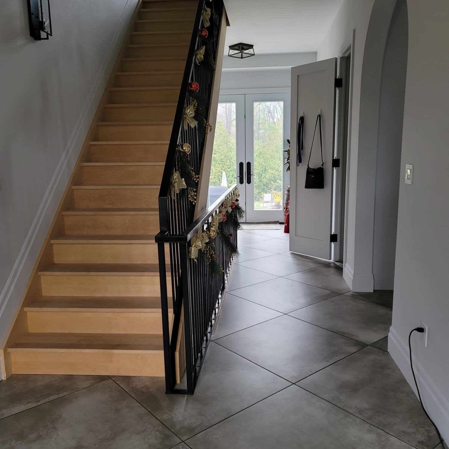 Jasne drewniane schody z czarną balustradą udekorowaną świątecznymi ozdobami. Szara podłoga z płytek ułożonych w romby. Białe drzwi i ściany. Nowoczesne wykończenie wnętrza.