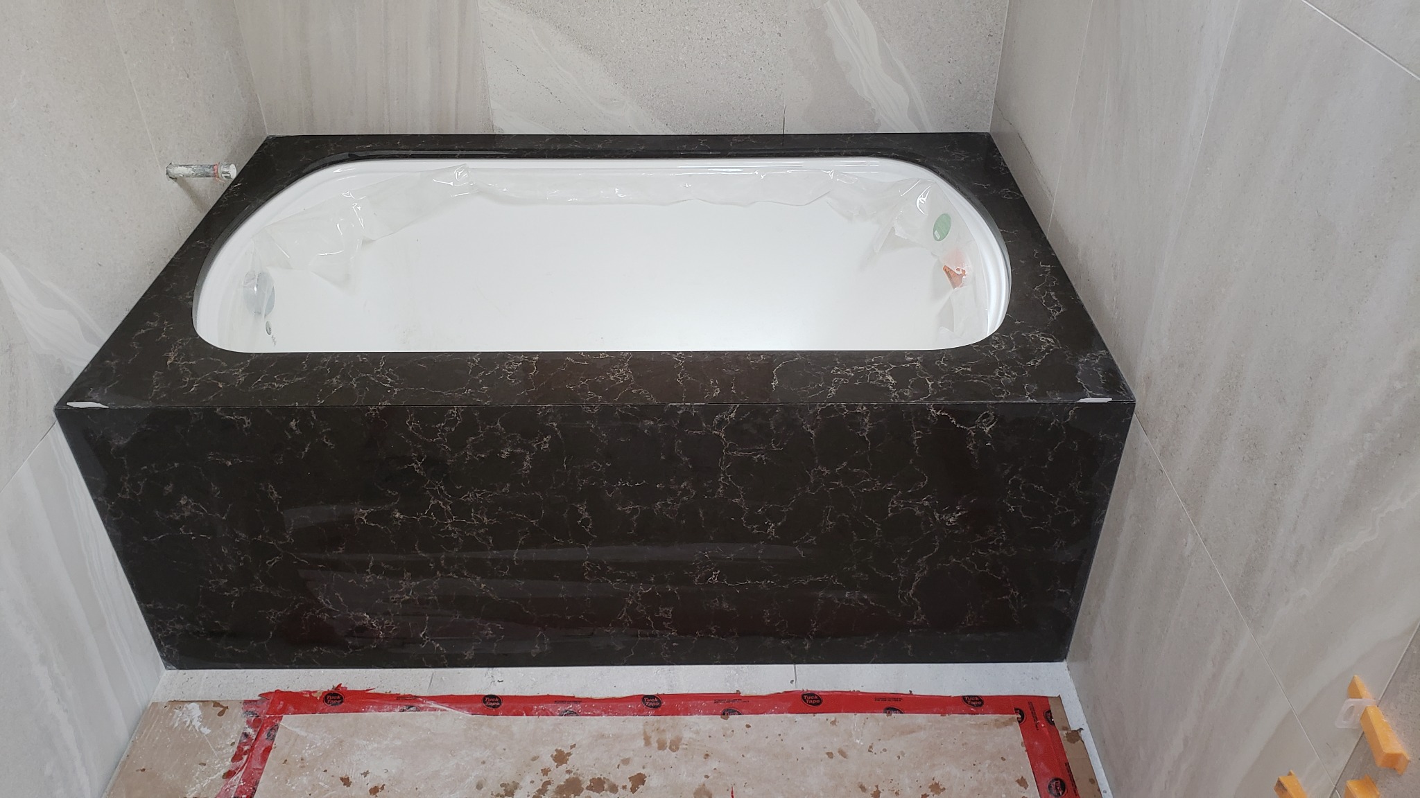 Obudowana wanna z ciemnego marmuru z białym wnętrzem, zabezpieczona folią. W tle jasne, marmurowe płytki i fragment podłogi w trakcie remontu.
