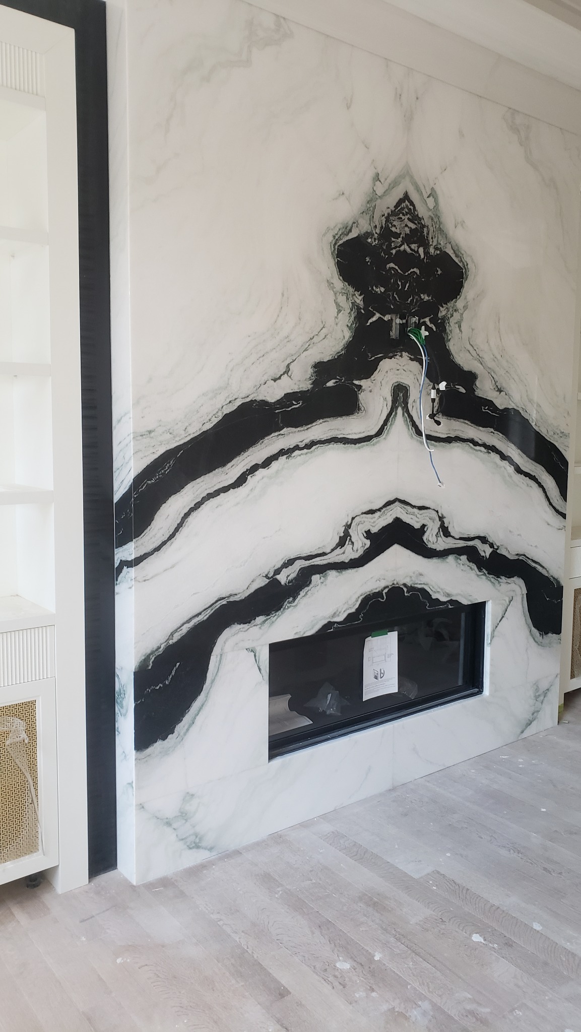 Ściana wykończona białym marmurem z czarnymi żyłami, wbudowany czarny wkład kominkowy, widoczne przewody elektryczne. Nowoczesne wykończenie wnętrza.