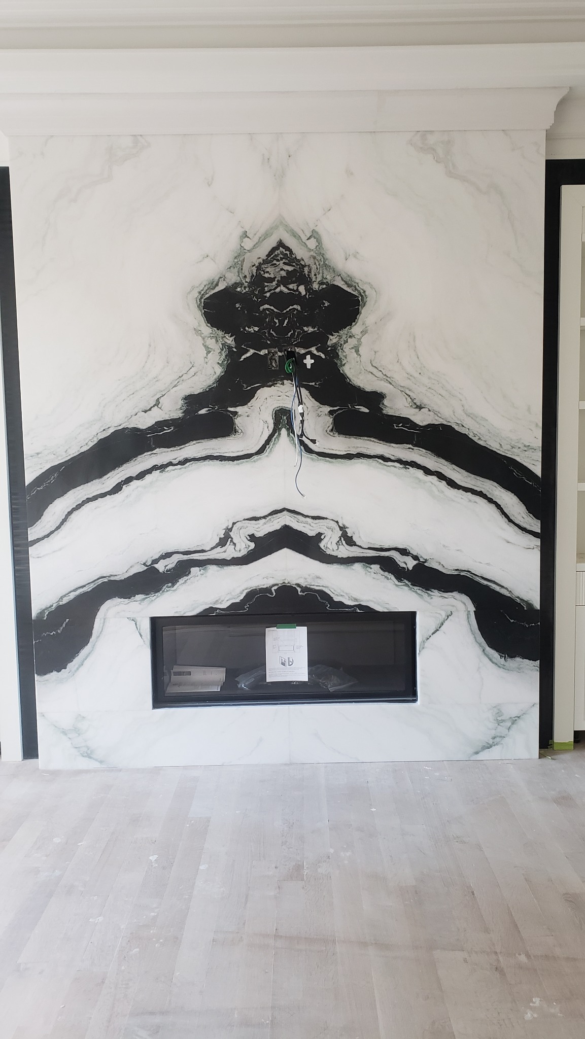 Ściana z marmurowymi płytami z czarnym wzorem nad kominkiem. Widoczne kable elektryczne. Całość w jasnej tonacji, podłoga drewniana.