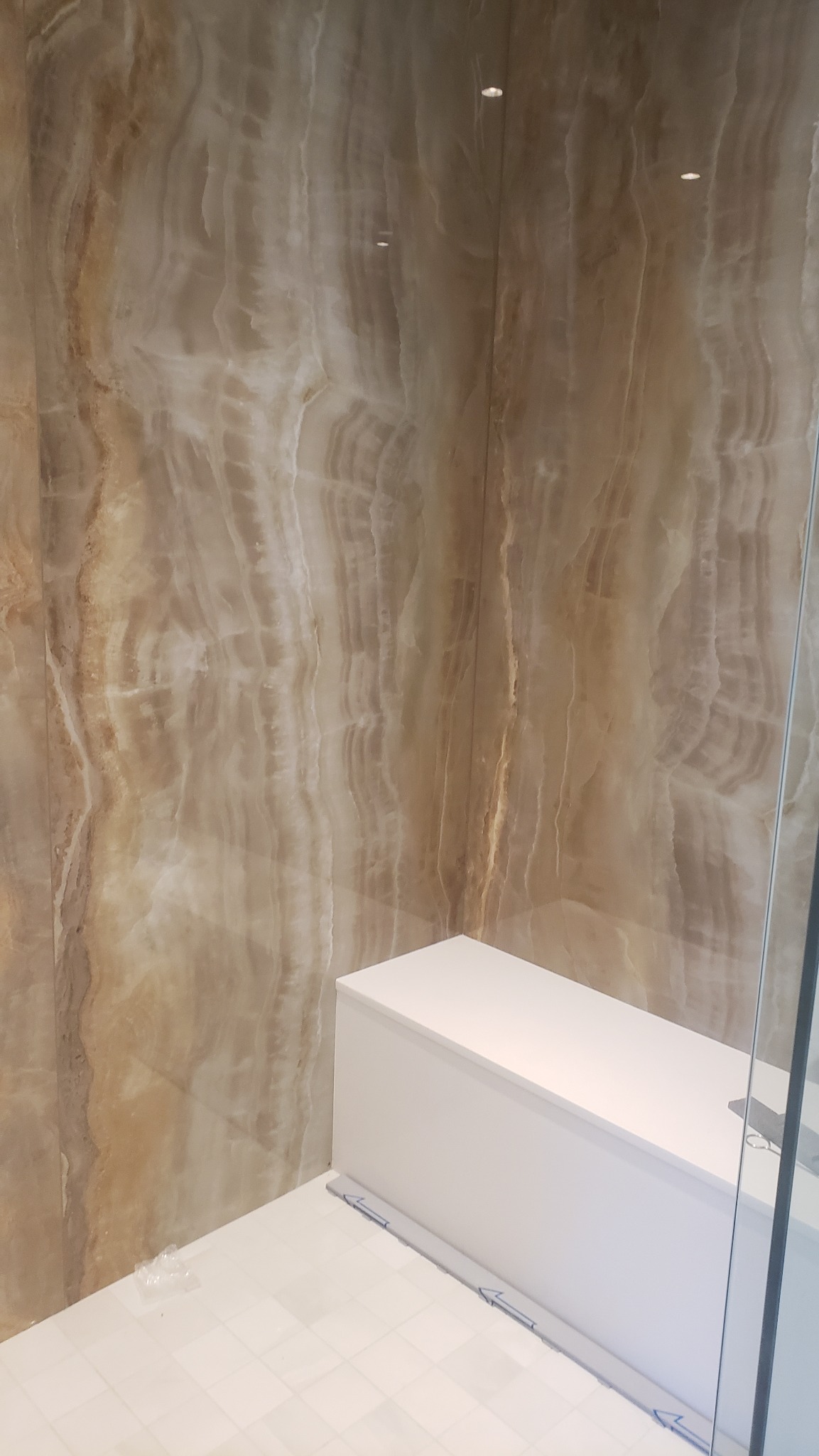 Wykończona łazienka z beżowymi płytkami imitującymi marmur na ścianach, białą zabudową i podłogą z jasnych płytek. Widoczny fragment kabiny prysznicowej.