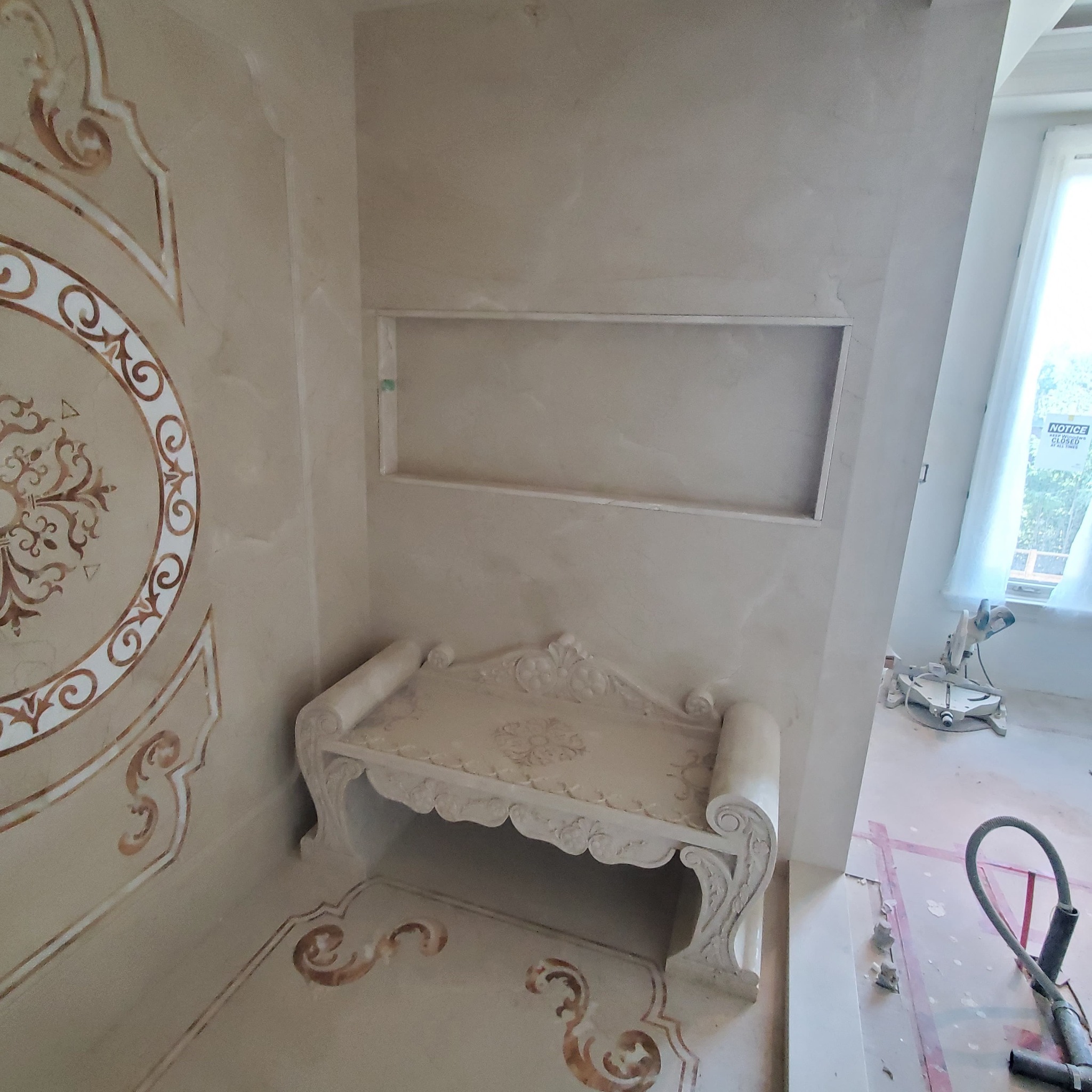 Wykończona ściana z dekoracyjnym ornamentem i wnęką, zdobiona ławka. W tle narzędzia sugerujące prace wykończeniowe.