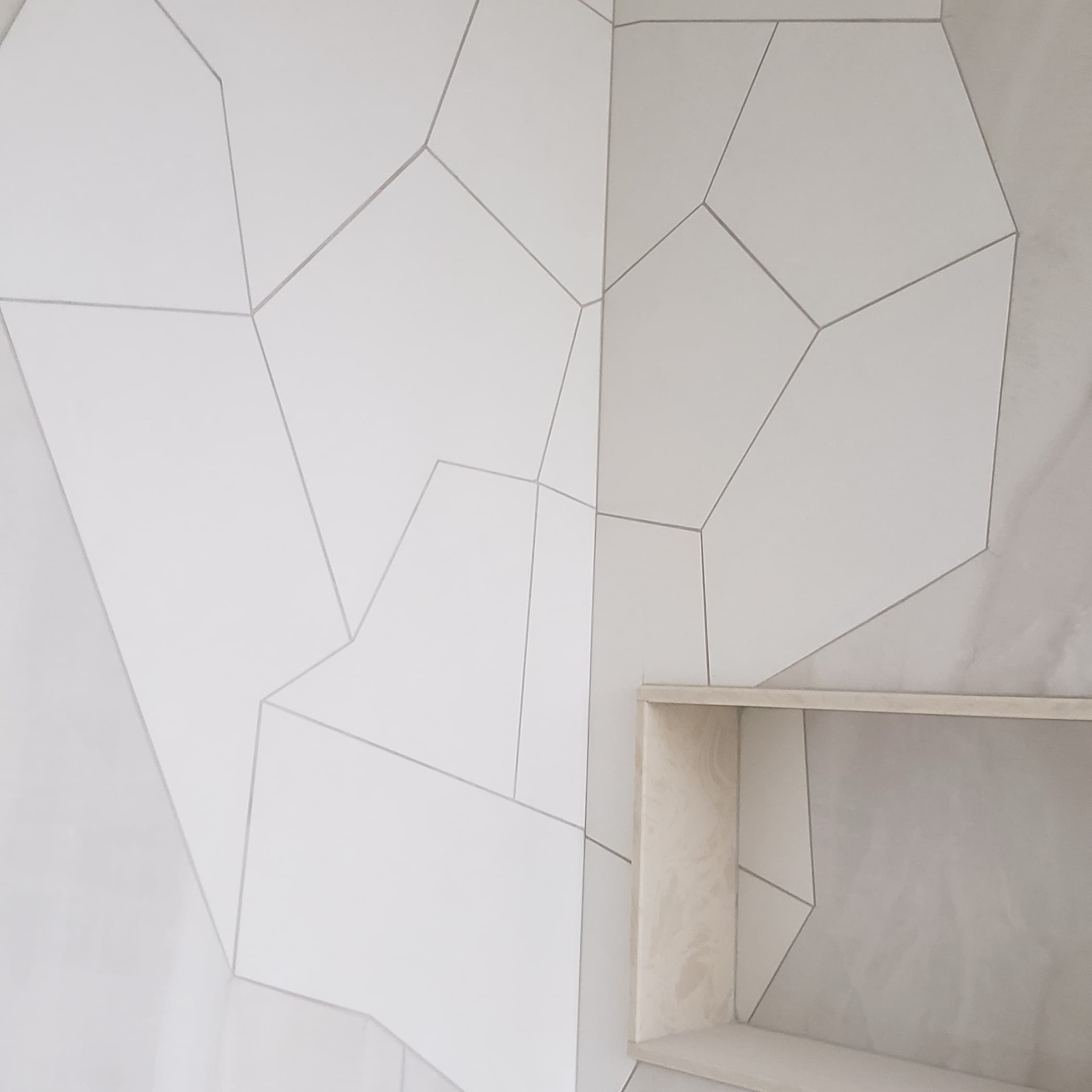 Ściana z białych, nieregularnych płytek z szarymi fugami i wnęką. Minimalistyczny design, neutralna kolorystyka. Częściowy widok na ścianę.