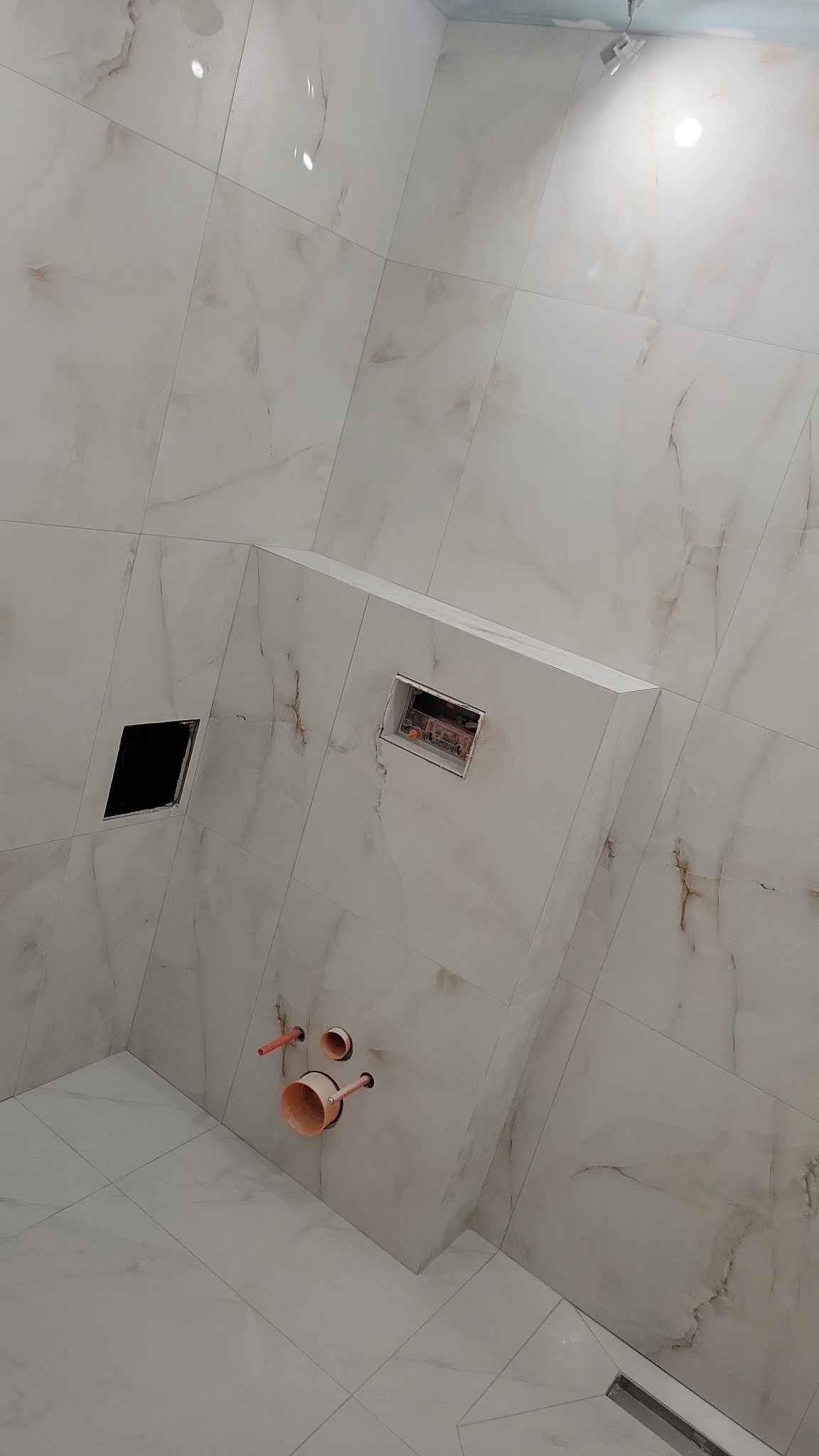 Wykończona łazienka z beżowymi płytkami imitującymi marmur. Widoczne otwory na instalacje sanitarne i odpływ liniowy w podłodze. Ujęcie z dołu, podkreślające przestrzeń.