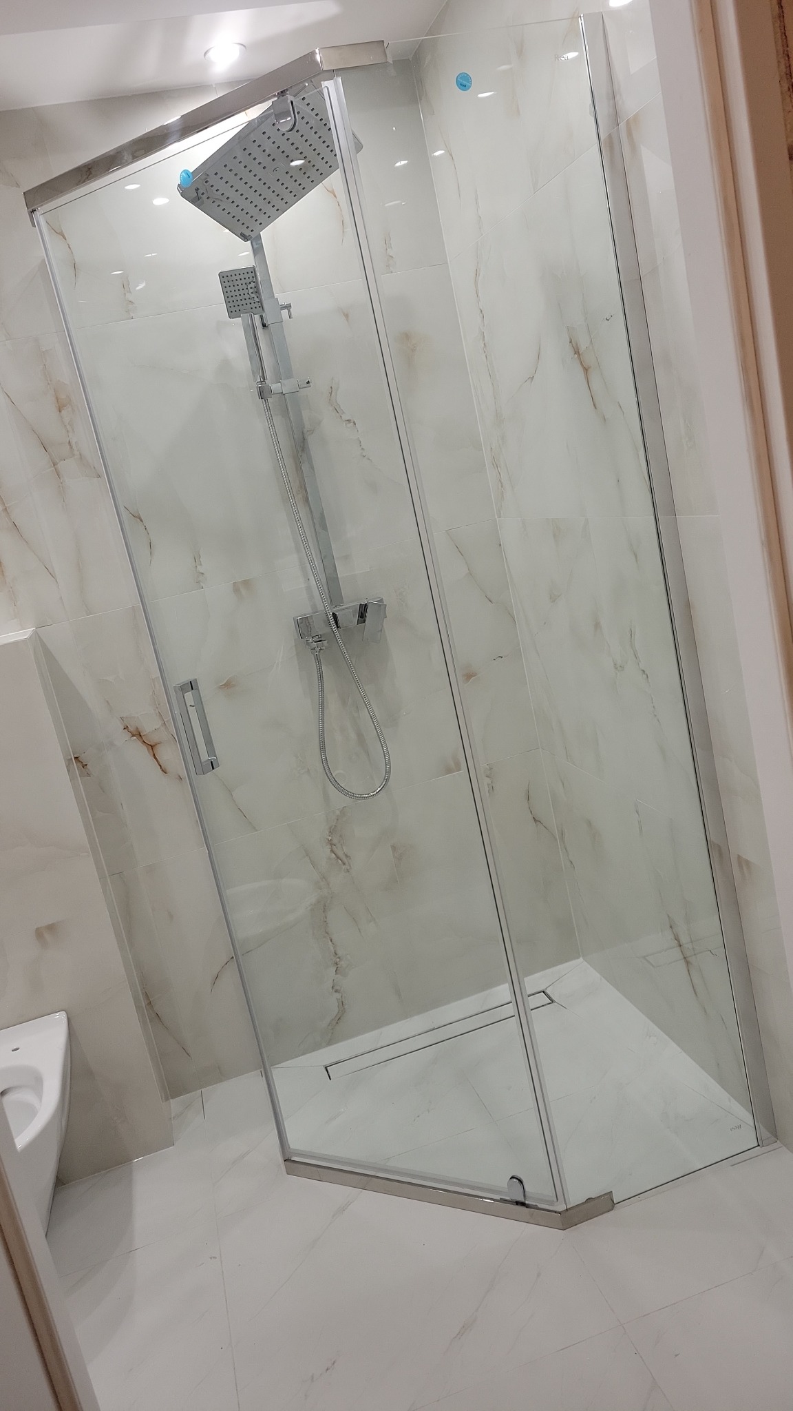 Nowoczesna kabina prysznicowa z deszczownicą, wykończona marmurowymi płytkami w odcieniach bieli i beżu. Widoczny odpływ liniowy w posadzce.