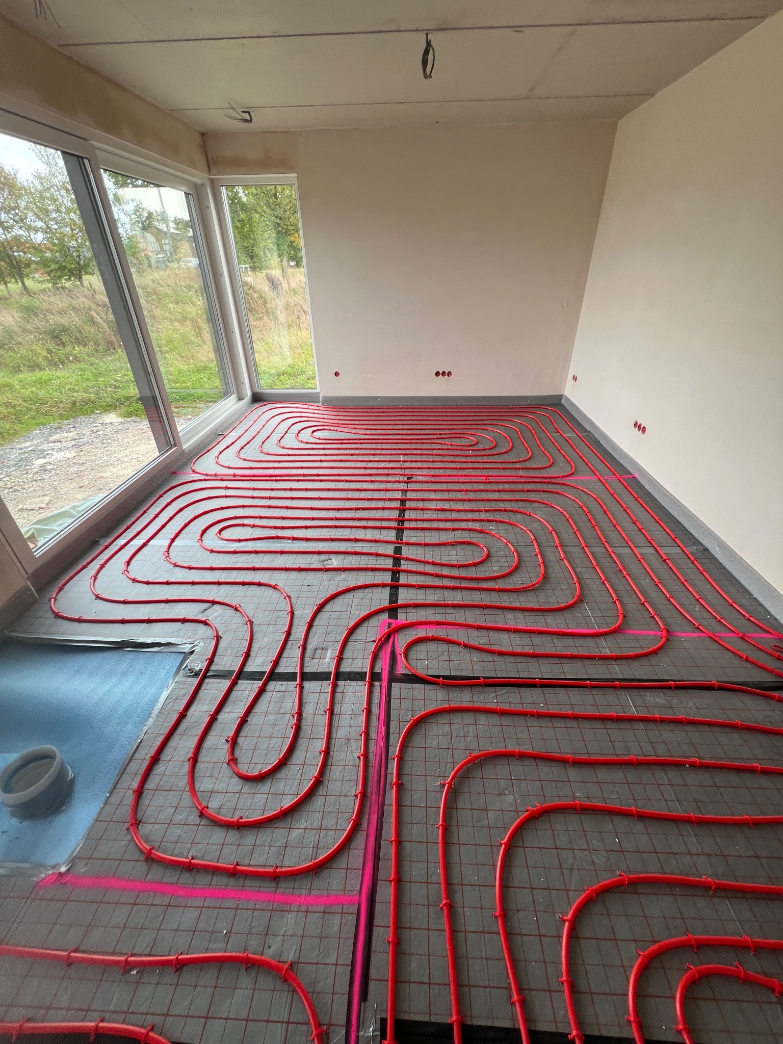 Czerwone rury ogrzewania podłogowego ułożone na szarej macie w jasnym pomieszczeniu z dużym oknem, widok z góry, etap instalacji.