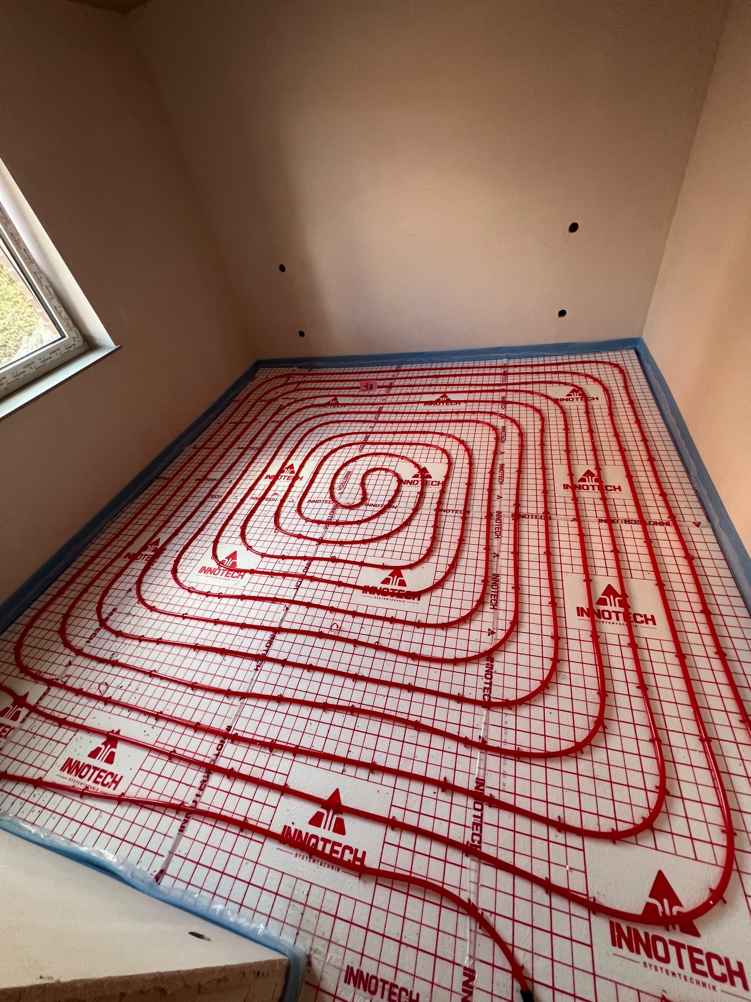Czerwone rury ogrzewania podłogowego Innotech ułożone spiralnie na białej macie z siatką w nowym pomieszczeniu z oknem na poddaszu, gotowe do zalania wylewką.