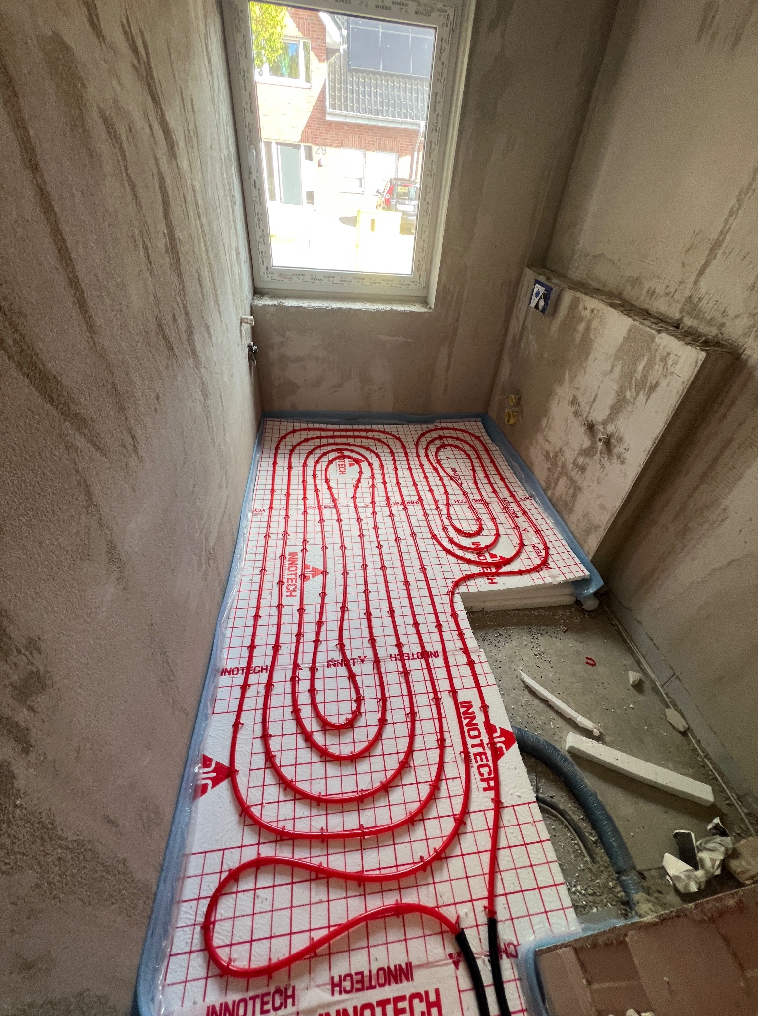 Układanie czerwonych rur ogrzewania podłogowego na białej macie izolacyjnej w surowym pomieszczeniu z oknem dachowym. Widoczne elementy instalacji i narzędzia.