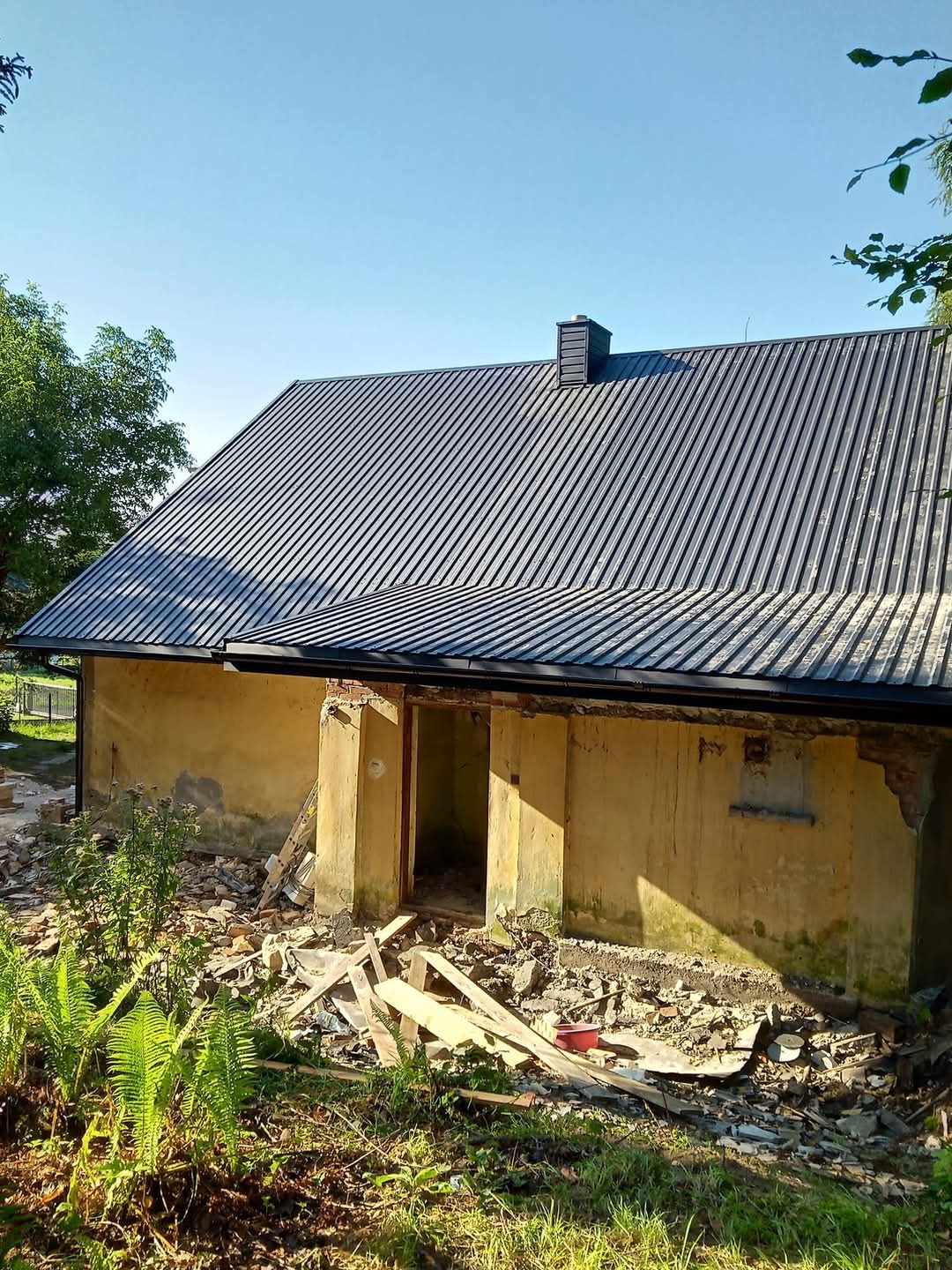 Budynek z nowym, ciemnoszarym dachem z blachy trapezowej. Wokół budynku gruz i pozostałości po remoncie. Widoczne zielone rośliny i fragment błękitnego nieba.