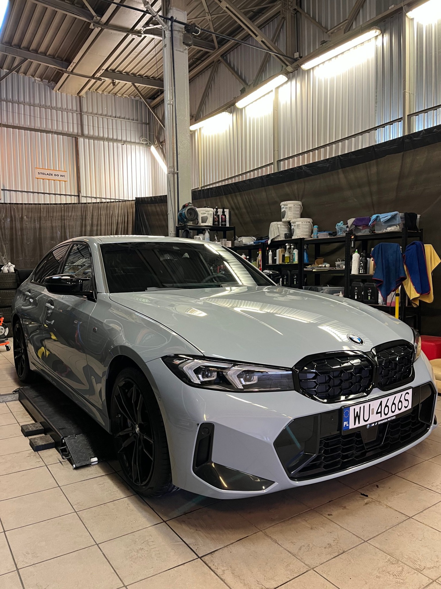 Szary samochód BMW serii 3 na podnośniku w warsztacie. Widoczne czarne felgi i tablica rejestracyjna. W tle regał z preparatami i oświetlenie warsztatowe.