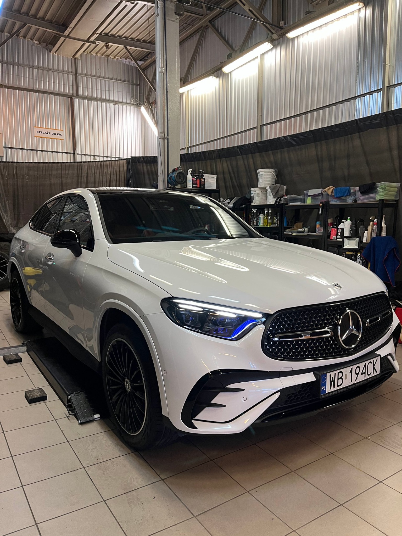 Biały Mercedes GLC Coupe na podnośniku w warsztacie, z widocznymi narzędziami i środkami czystości na półkach w tle. Auto gotowe do prania tapicerki.