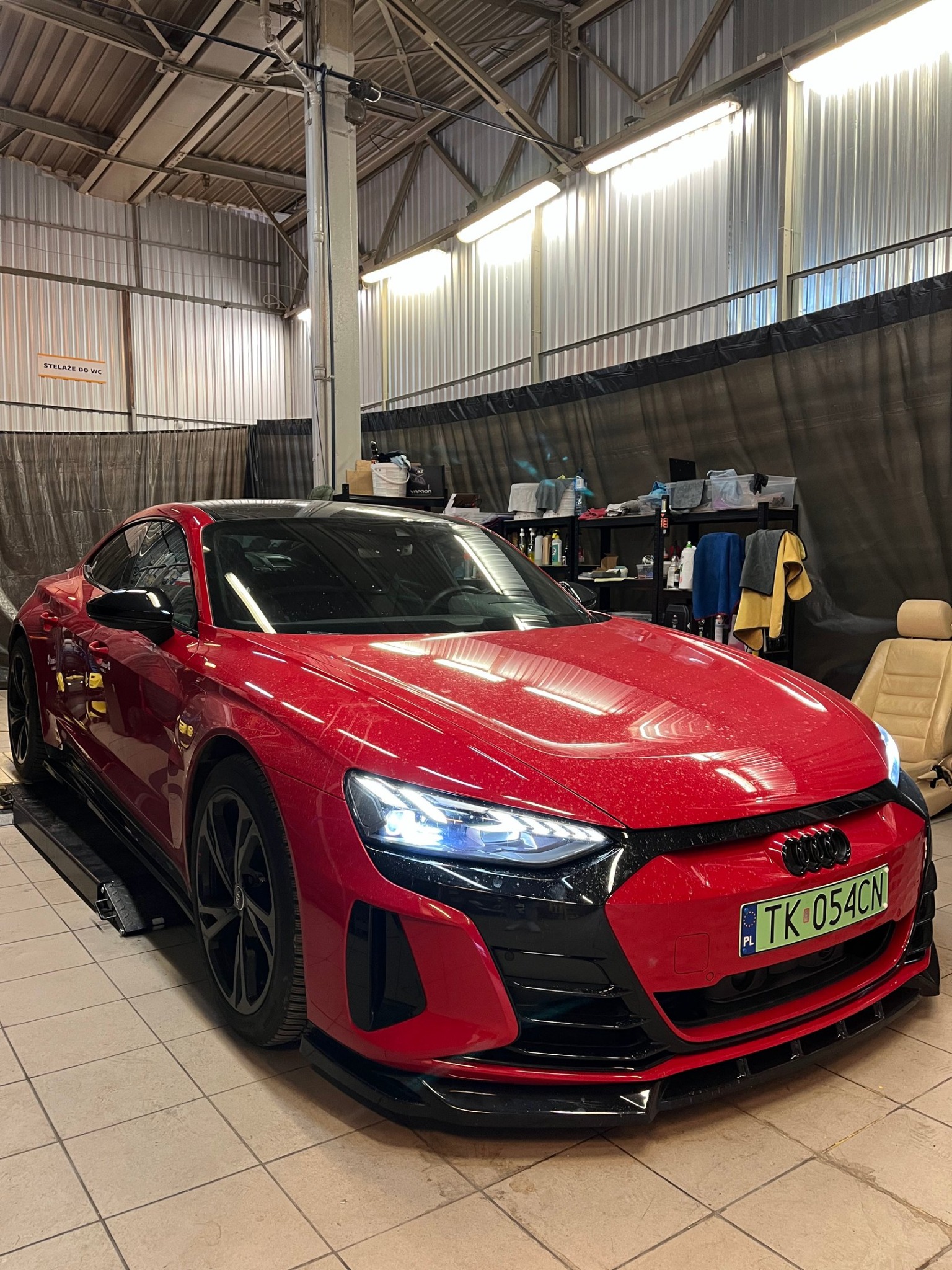 Czerwone Audi e-tron GT na podnośniku w warsztacie, widoczne zabrudzenia na karoserii, fotel samochodowy obok, oświetlenie warsztatowe.