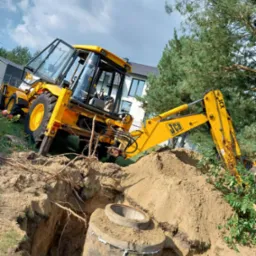 Żółta koparko-ładowarka JCB przy wykopie z betonową rurą, drzewa w tle. Prace ziemne w toku, widoczne korzenie i świeża ziemia.