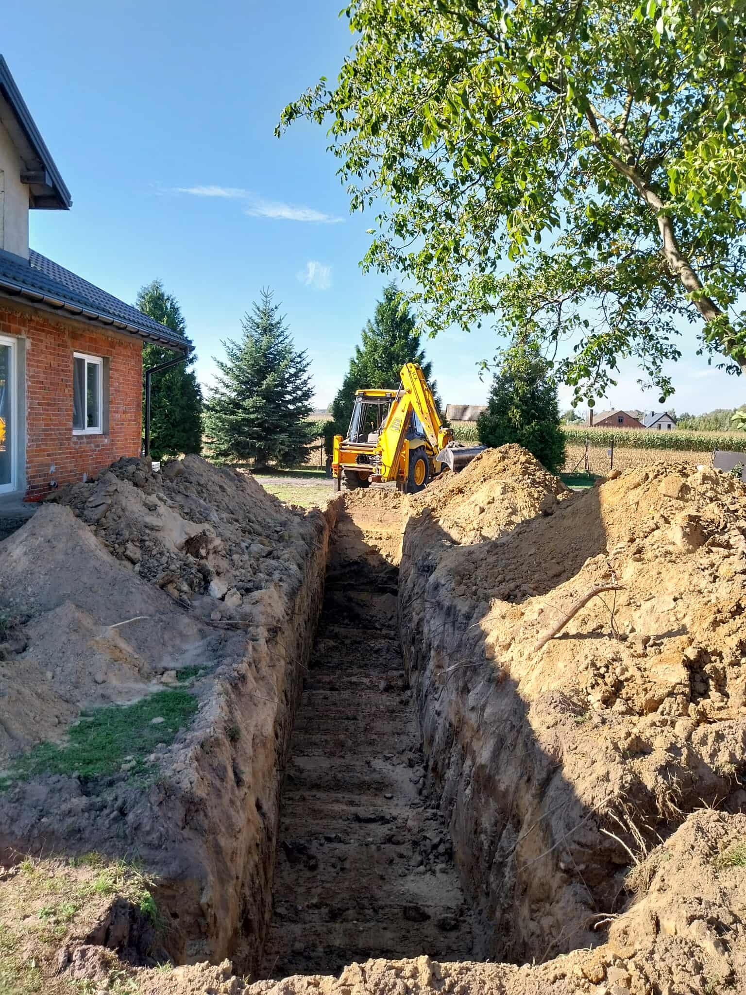Żółta koparko-ładowarka JCB obok wykopu liniowego w ziemi, widok z dołu. W tle dom z czerwonej cegły i zielone drzewa pod błękitnym niebem.
