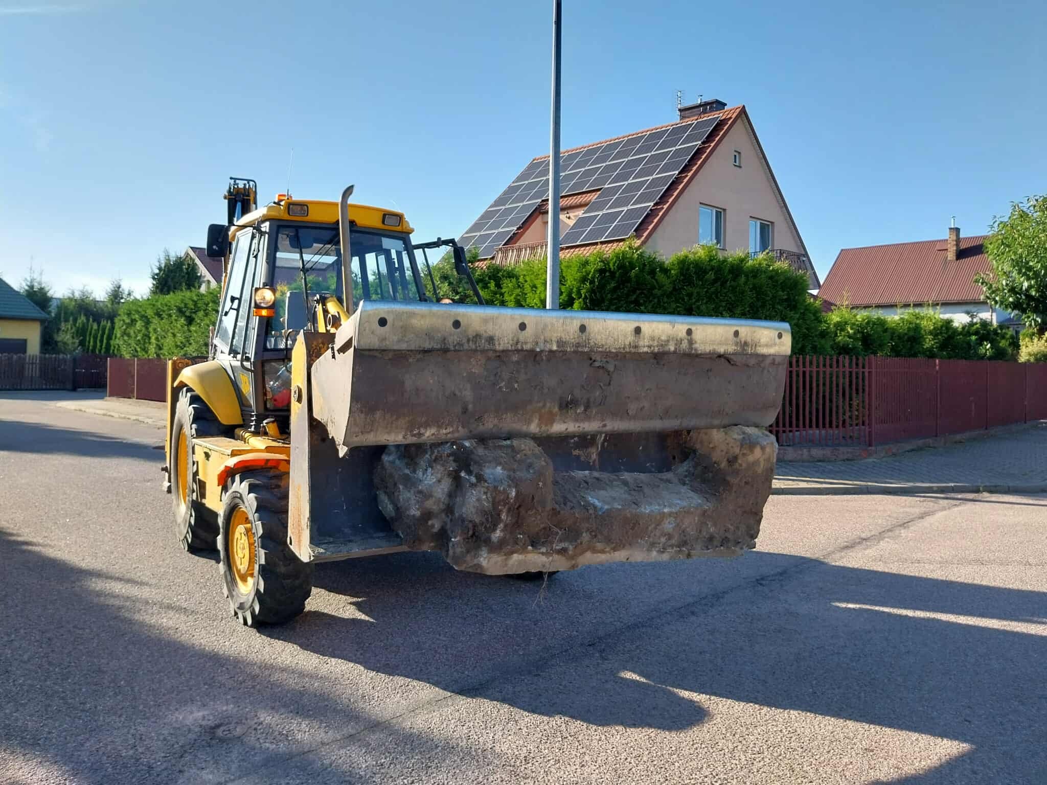 Żółta koparko-ładowarka z pełną łyżką ziemi na tle domów z panelami słonecznymi, na osiedlowej drodze. Ujęcie z bliska, widok z przodu, słoneczny dzień.