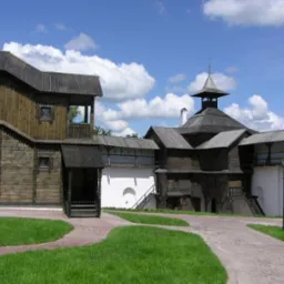 Kompleks budynków z ciemnego drewna, w stylu tradycyjnej architektury rosyjskiej, połączonych białymi murami, z wieżą i zielonym trawnikiem na pierwszym planie.