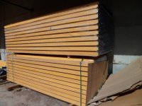 Stos tarcicy budowlanej w magazynie, widoczne deski o jasnym odcieniu drewna ułożone warstwami, część desek owinięta czarną folią.