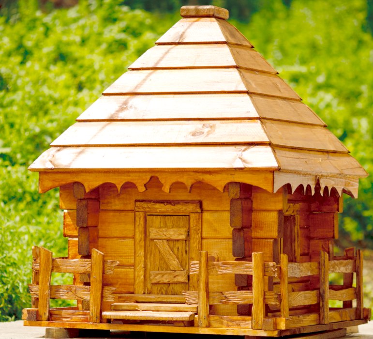 Mała, drewniana chatka z gontowym dachem i miniaturowym płotkiem, prawdopodobnie makieta lub domek dla lalek, ustawiona na tle zieleni.