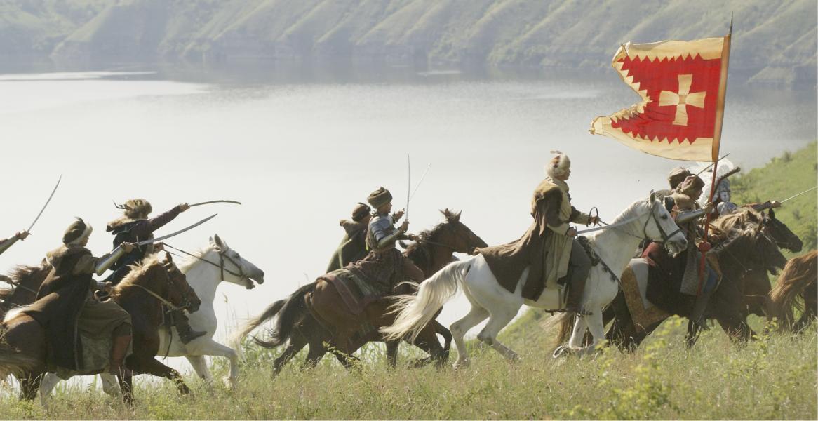 Grupa jeźdźców konnych w strojach historycznych z szablami i chorągwią z krzyżem na tle jeziora i wzgórz.