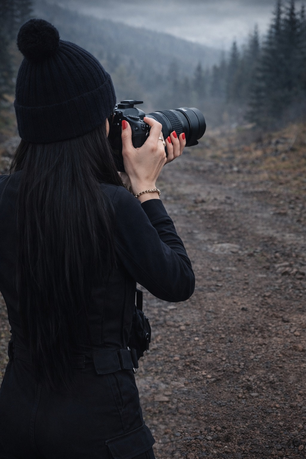 Kobieta w czarnym kombinezonie i czapce z pomponem robi zdjęcie aparatem z teleobiektywem na tle zalesionych gór we mgle. Czerwone paznokcie i złota bransoletka.