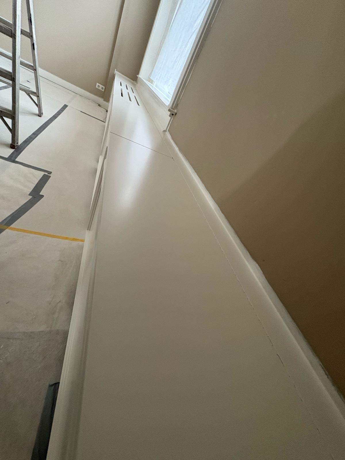 Świeżo pomalowana ściana z listwą przypodłogową, oklejone okno folią ochronną, drabina w rogu. Podłoga zabezpieczona taśmą malarską.
