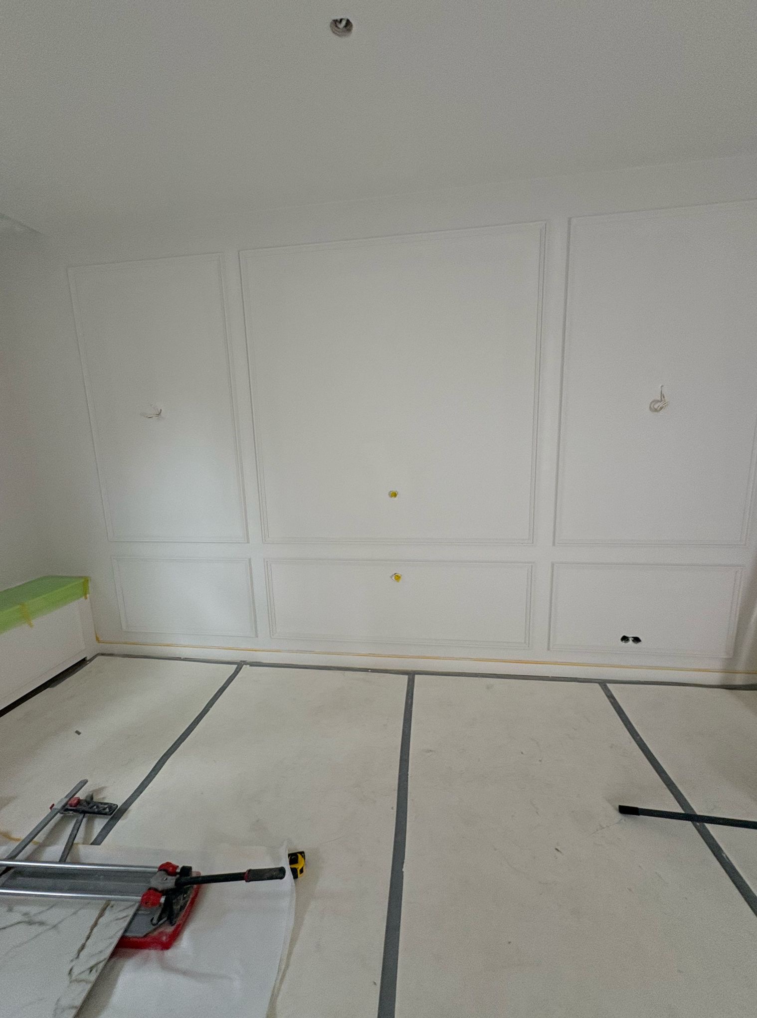 Białe ściany z dekoracyjnymi panelami i listwami w trakcie wykańczania. Na podłodze zabezpieczona powierzchnia i narzędzia do cięcia płytek.