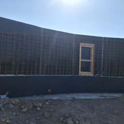 ProBud Adam Bąk - Szare szalunki fundamentowe z metalową siatką i drewnianą ramą okienną na tle jasnego nieba. Widoczny fragment gruntu z kamieniami i wystającymi elementami zbrojenia.