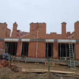 Budowa domu z czerwonej cegły Porotherm w Rybniku, widoczne okna i rusztowania. Prace murarskie w trakcie realizacji, cegły ułożone do wysokości okien.