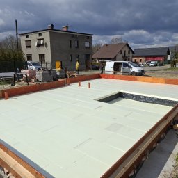 ProBud Adam Bąk - Przygotowany fundament z izolacją termiczną, obudowany szalunkiem. Widoczne wystające rury wentylacyjne. W tle budynki mieszkalne i pochmurne niebo.