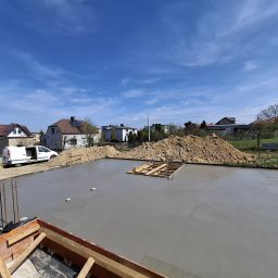 ProBud Adam Bąk - Świeżo wylany, gładki fundament pod budowę domu, z drewnianym szalunkiem pośrodku, pod błękitnym niebem. W tle zabudowa jednorodzinna i zaparkowany biały bus.