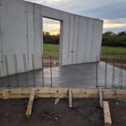 ProBud Adam Bąk - Świeżo wylany beton na fundamencie z wystającymi prętami zbrojeniowymi i drewnianym szalunkiem. W tle widoczne pole i zachodzące słońce.