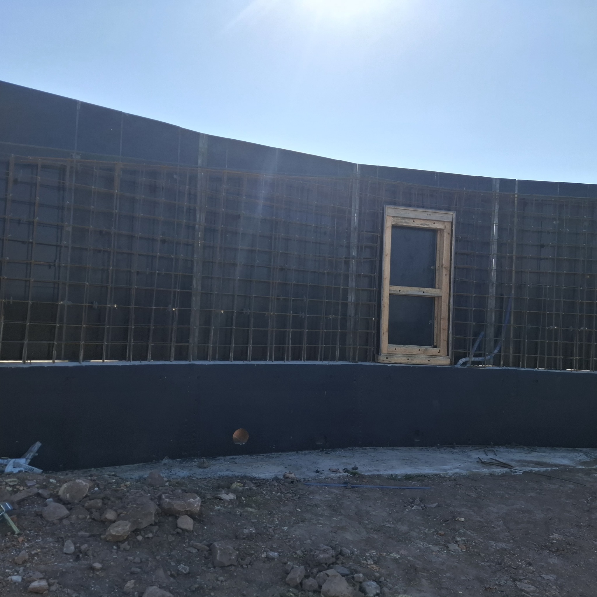 Szare szalunki fundamentowe z metalową siatką i drewnianą ramą okienną na tle jasnego nieba. Widoczny fragment gruntu z kamieniami i wystającymi elementami zbrojenia.