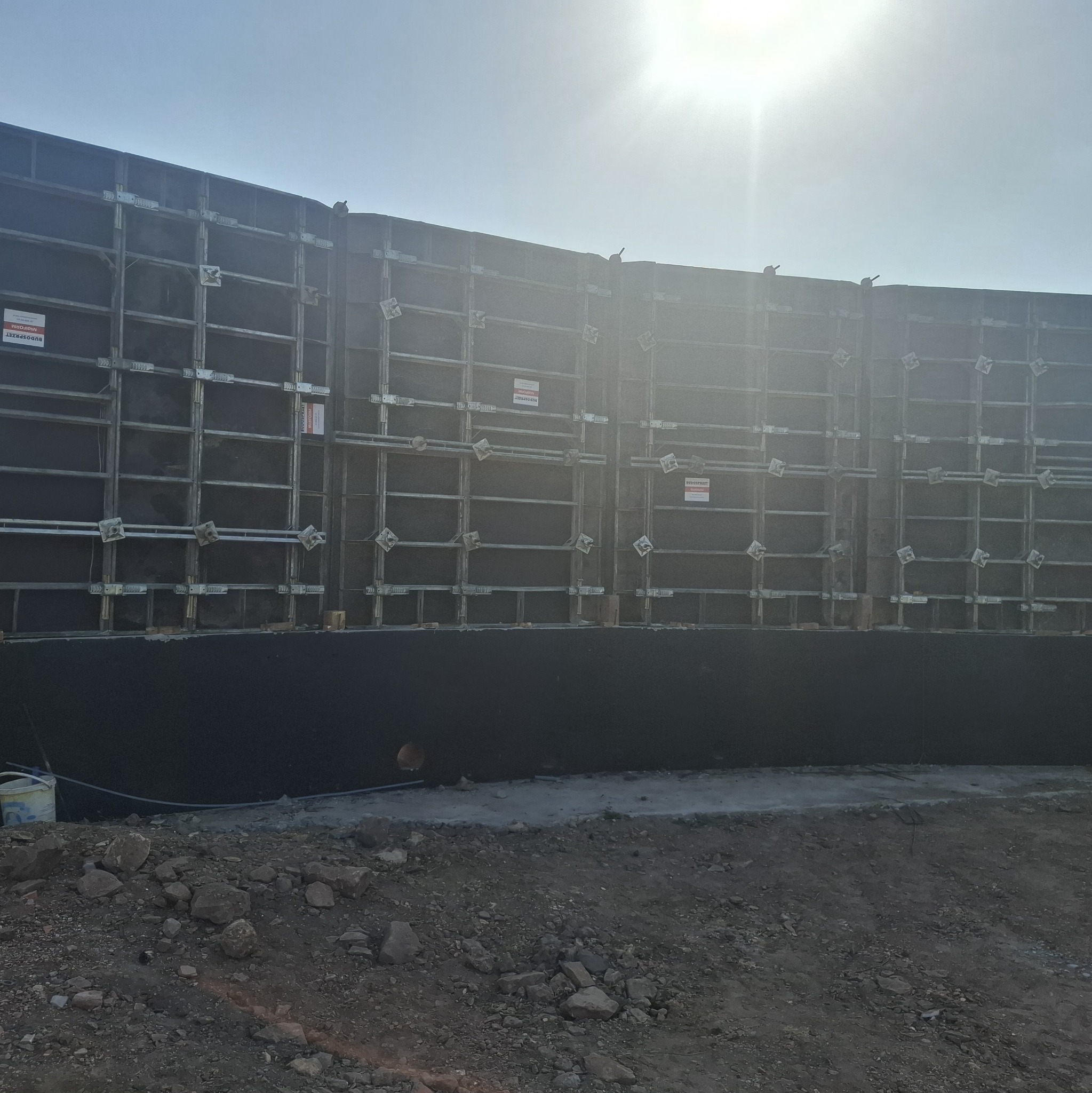 Szare szalunki z metalową konstrukcją na budowie, w tle jasne niebo i słońce. Widoczne tabliczki informacyjne na szalunkach.