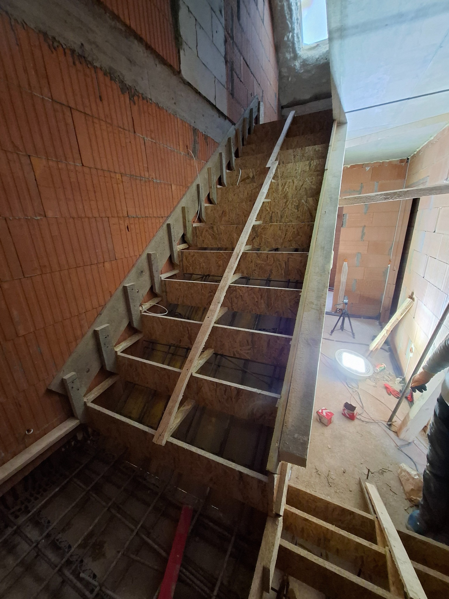 Szkielet drewnianych schodów z płyt OSB w surowym stanie, widoczne zbrojenie w podłożu, ceglane ściany. Budowa w trakcie realizacji.