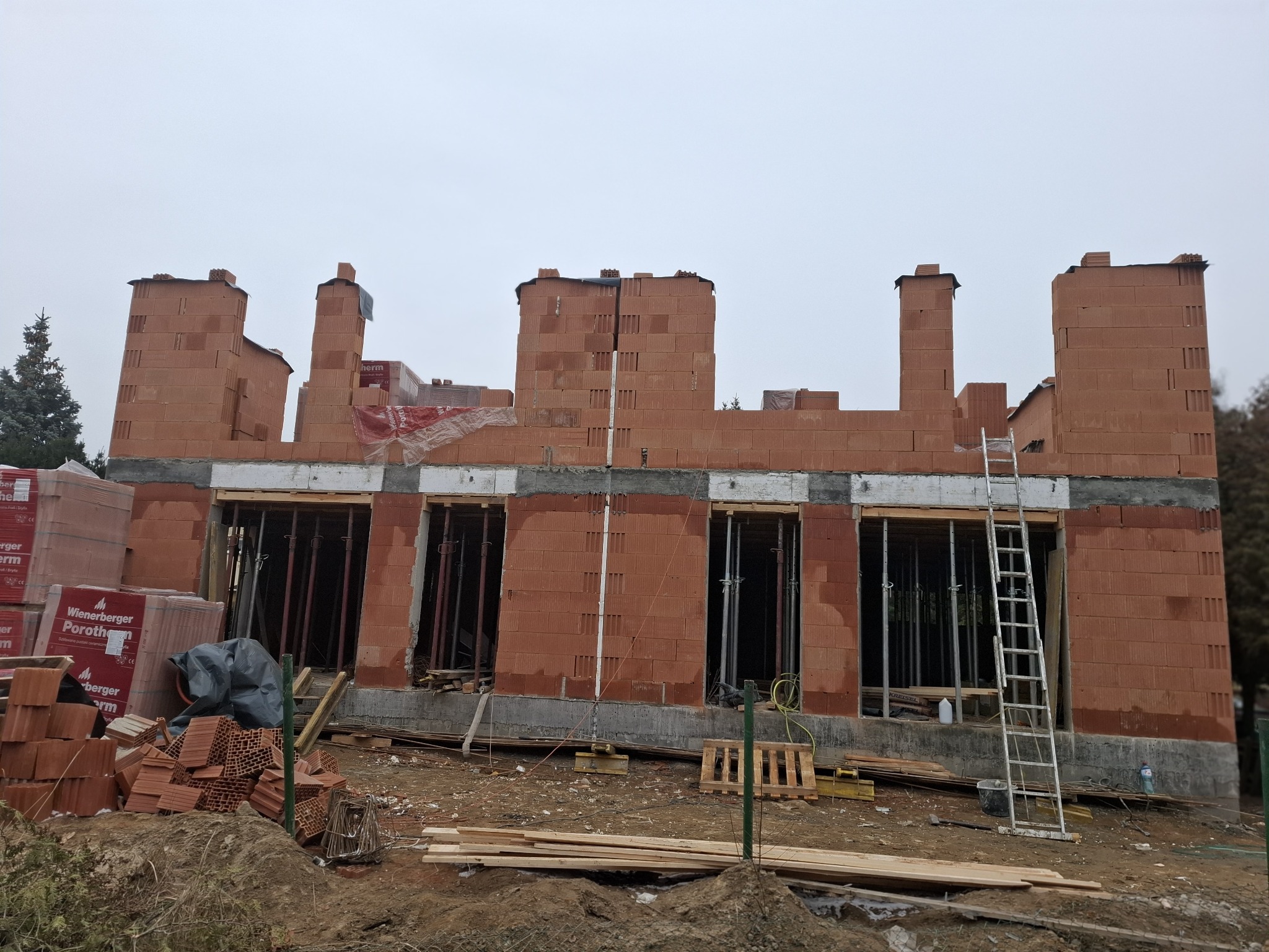 Budowa domu z czerwonej cegły Porotherm w Rybniku, widoczne okna i rusztowania. Prace murarskie w trakcie realizacji, cegły ułożone do wysokości okien.