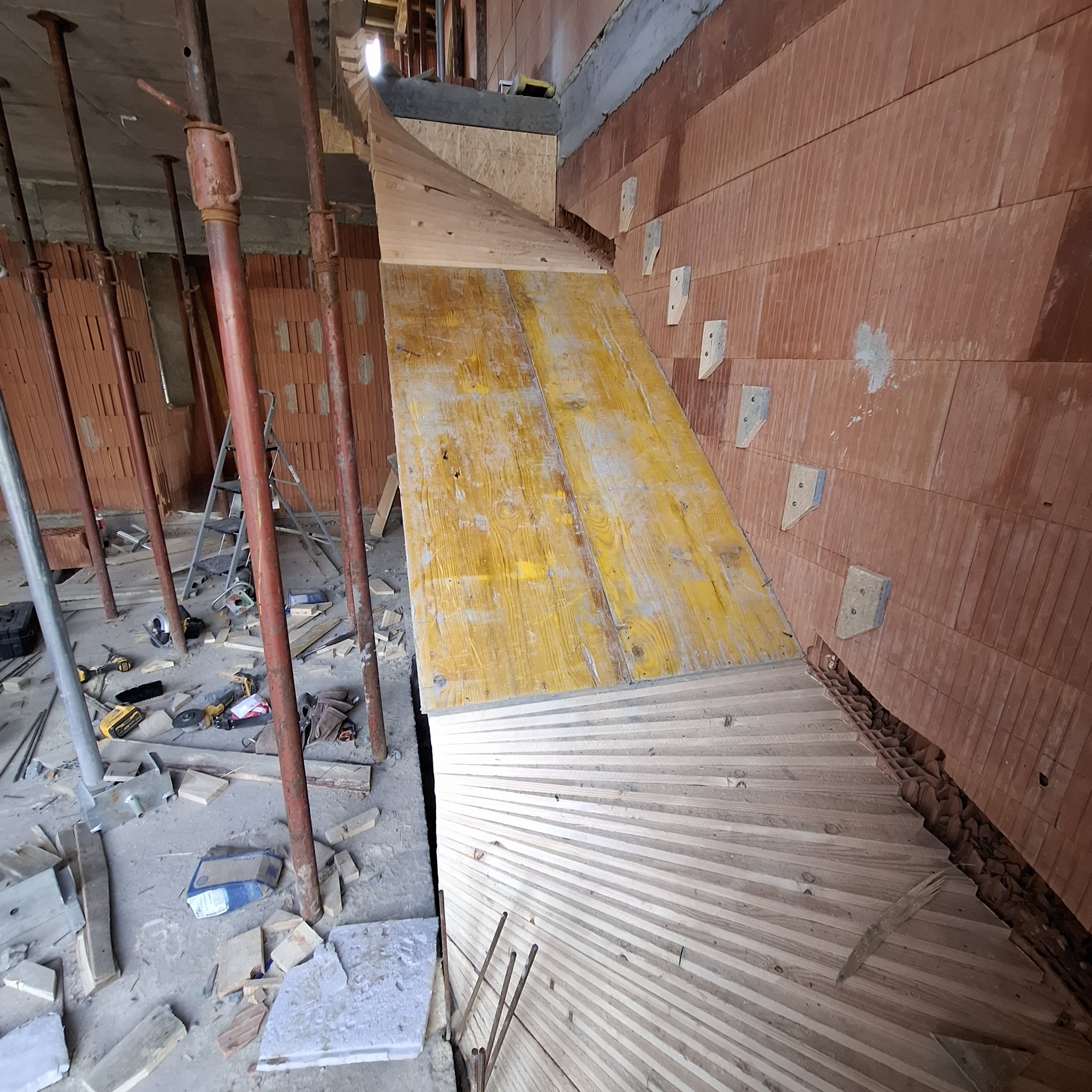 Murowana ściana z elementami szalunku i stemplami. Widoczne narzędzia i materiały budowlane. Kąt ujęcia z dołu podkreśla wysokość konstrukcji.