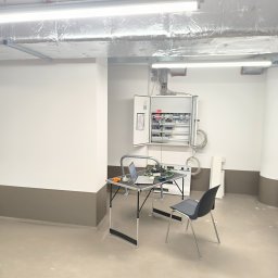 ENCEL ELECTRO SPÓŁKA Z OGRANICZONĄ ODPOWIEDZIALNOŚCIĄ - Otwarta skrzynka z instalacją elektryczną w pomieszczeniu. Na stole laptop i narzędzia, krzesło obok. Widoczne rury wentylacyjne pod sufitem.