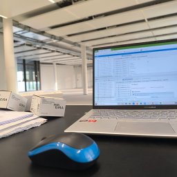 ENCEL ELECTRO SPÓŁKA Z OGRANICZONĄ ODPOWIEDZIALNOŚCIĄ - Laptop z oprogramowaniem KNX, mysz i pudełka GIRA na biurku. W tle widoczne przestrzenne, jasne wnętrze biurowe z oświetleniem sufitowym.