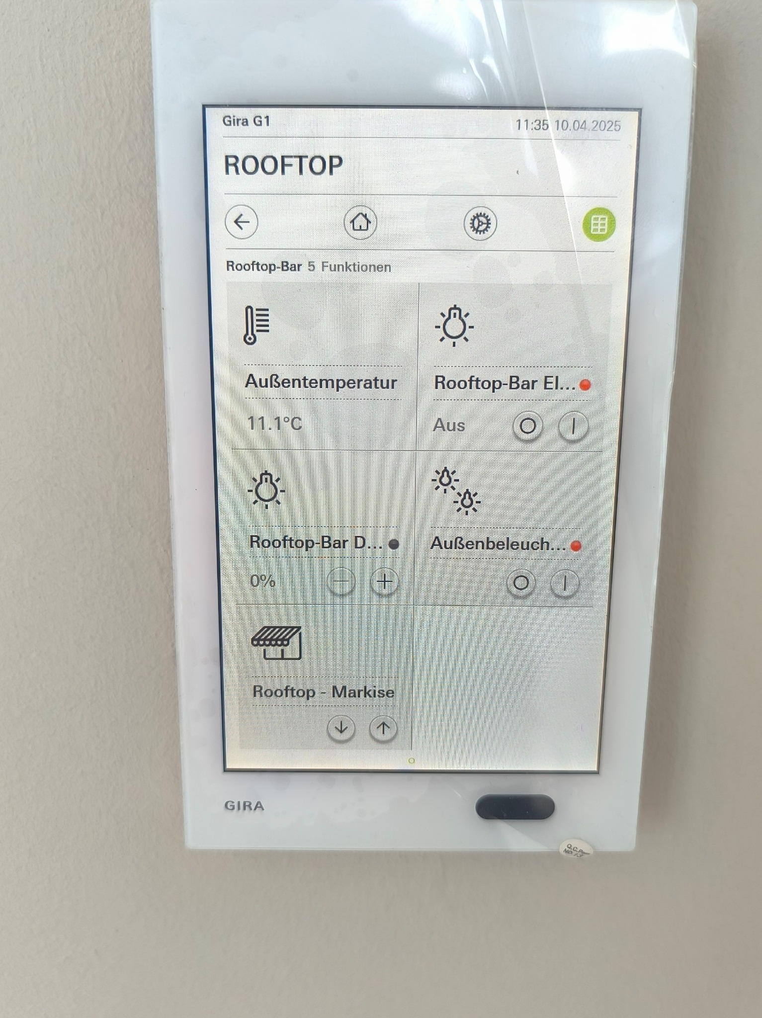 Biały panel dotykowy Gira G1 na ścianie, z interfejsem sterowania inteligentnym domem: temperatura, oświetlenie zewnętrzne i markiza na dachu. Widoczne opcje sterowania.