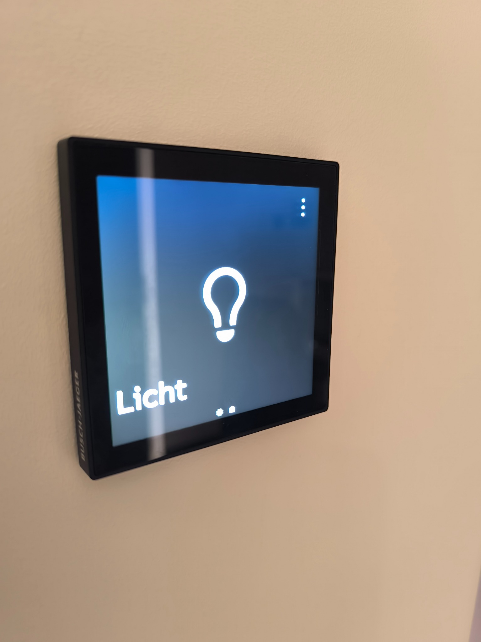 Panel dotykowy Busch-Jaeger zamontowany na ścianie, sterujący oświetleniem, z ikoną żarówki i napisem 'Licht' na niebieskim tle.