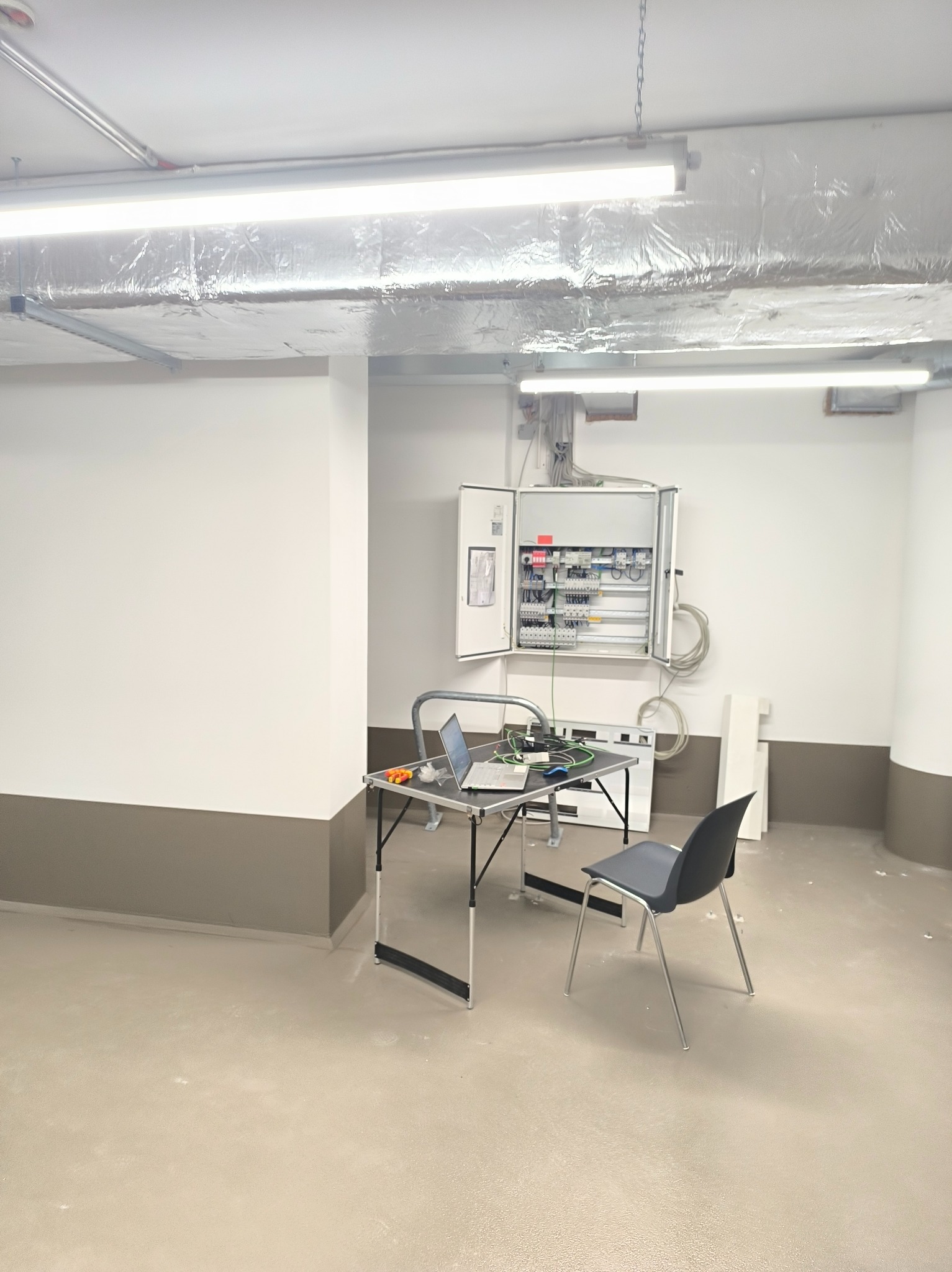 Otwarta skrzynka z instalacją elektryczną w pomieszczeniu. Na stole laptop i narzędzia, krzesło obok. Widoczne rury wentylacyjne pod sufitem.