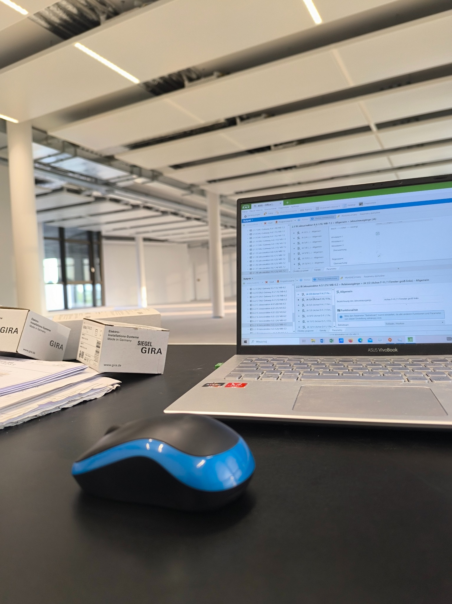 Laptop z oprogramowaniem KNX, mysz i pudełka GIRA na biurku. W tle widoczne przestrzenne, jasne wnętrze biurowe z oświetleniem sufitowym.