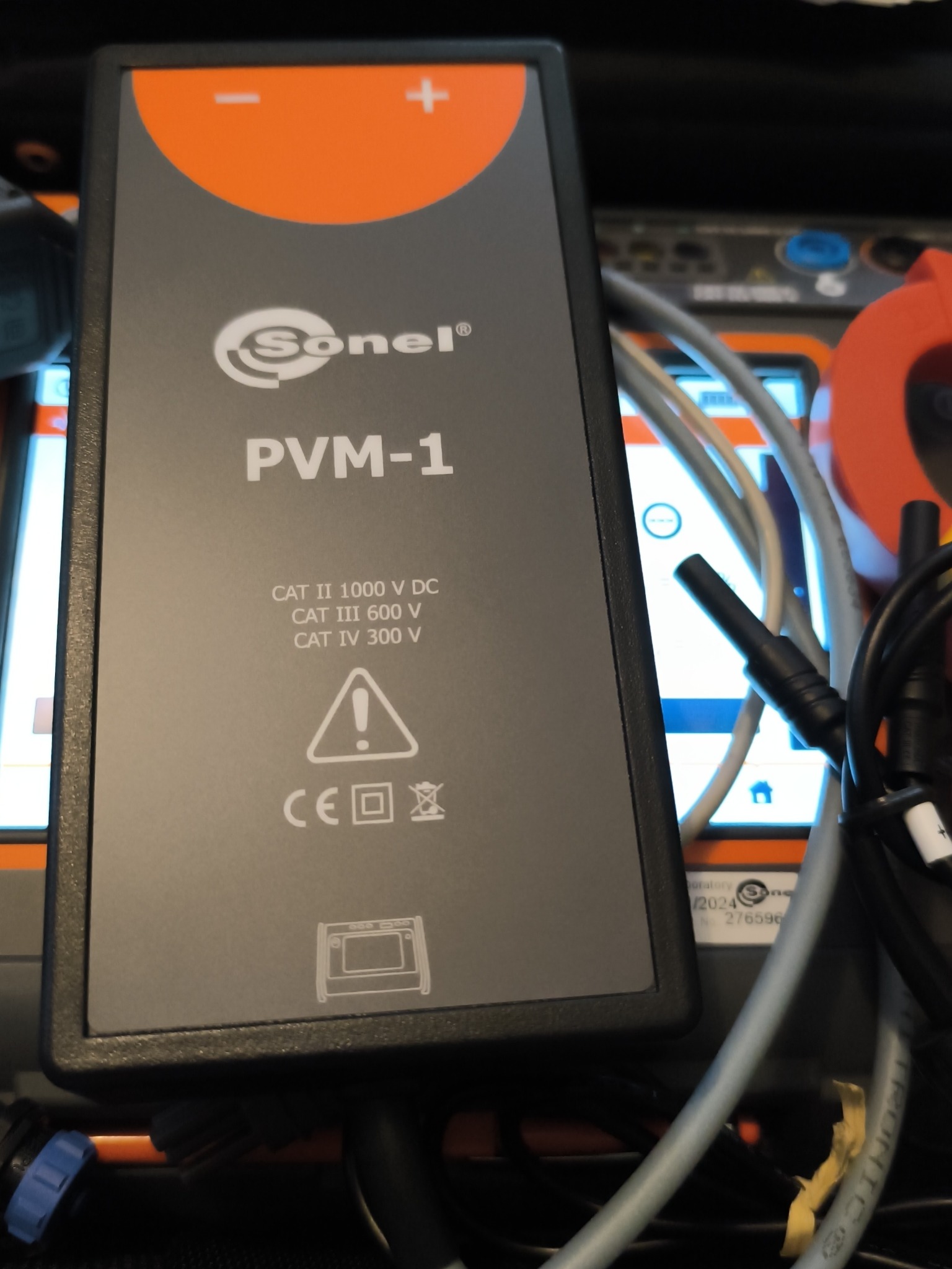 Miernik Sonel PVM-1 do pomiarów elektrycznych, z widocznymi kablami i fragmentami innych urządzeń pomiarowych w tle. Urządzenie w obudowie z pomarańczowymi akcentami.