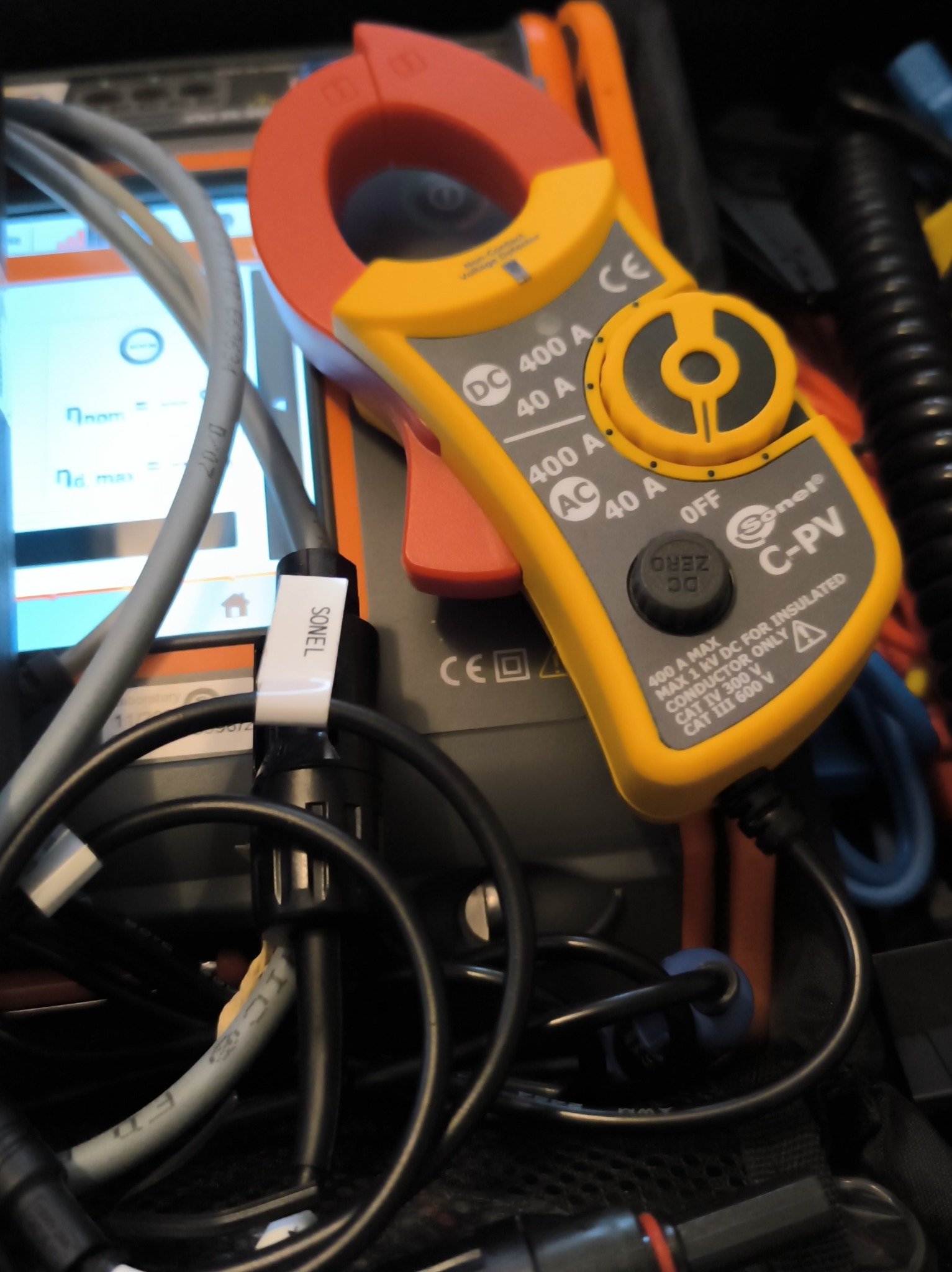 Żółto-czerwony miernik cęgowy Sonel C-PV na tle ekranu urządzenia pomiarowego i przewodów. Widoczne oznaczenia napięcia i prądu.