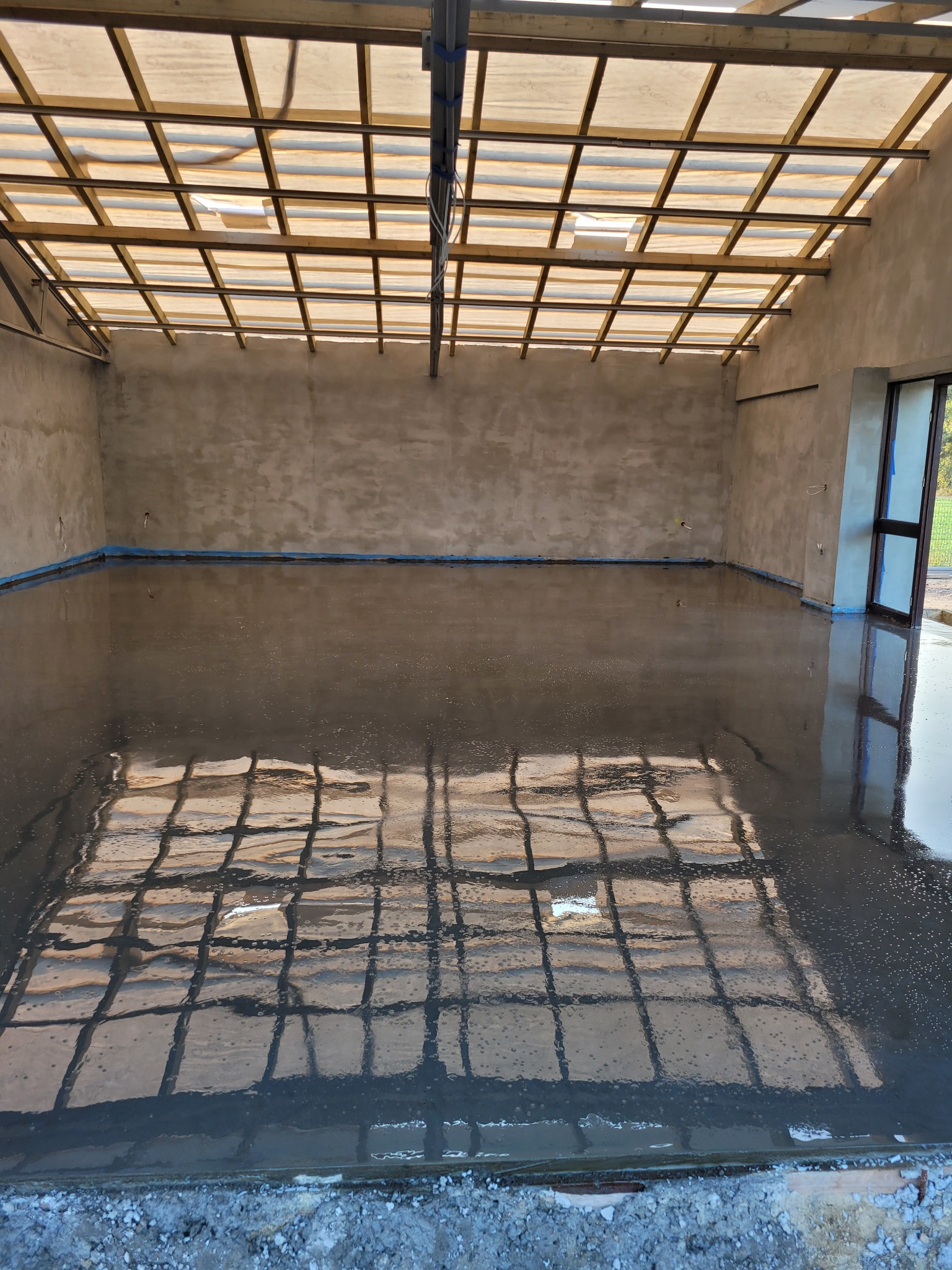 Świeżo wylana, gładka wylewka w pomieszczeniu z drewnianą konstrukcją dachu, zabezpieczona folią przy ścianach. Widoczne odbicie konstrukcji w mokrej powierzchni.