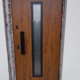 Brązowe drzwi zewnętrzne z pionowym przeszkleniem i czarną klamką, osadzone w surowym otworze muru z widocznym betonem i pianką montażową. Widok z bliska na zamontowane drzwi.