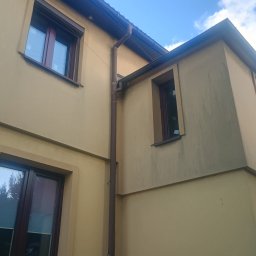 BB OKNA - MARCIN REMIZOW DAWID KRUPICZ SPÓŁKA CYWILNA - Fragment elewacji domu z brązowymi oknami i rynną. Widok z dołu na żółtą ścianę i błękitne niebo. Okna mają widoczne rolety zwinięte u góry.