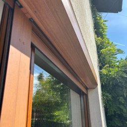 BB OKNA - MARCIN REMIZOW DAWID KRUPICZ SPÓŁKA CYWILNA - Drewnopodobne okno z częściowo opuszczoną roletą zewnętrzną, widok z dołu. Roleta w kolorze zbliżonym do ramy okiennej, na tle zieleni i jasnego nieba.