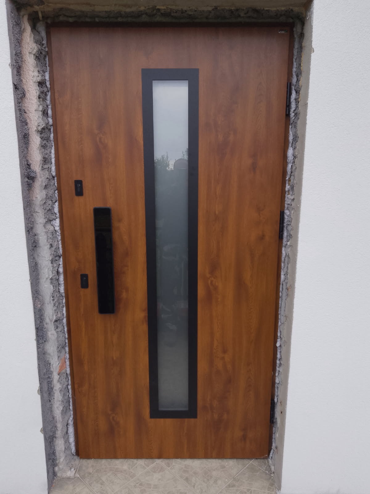 Brązowe drzwi zewnętrzne z pionowym przeszkleniem i czarną klamką, osadzone w surowym otworze muru z widocznym betonem i pianką montażową. Widok z bliska na zamontowane drzwi.