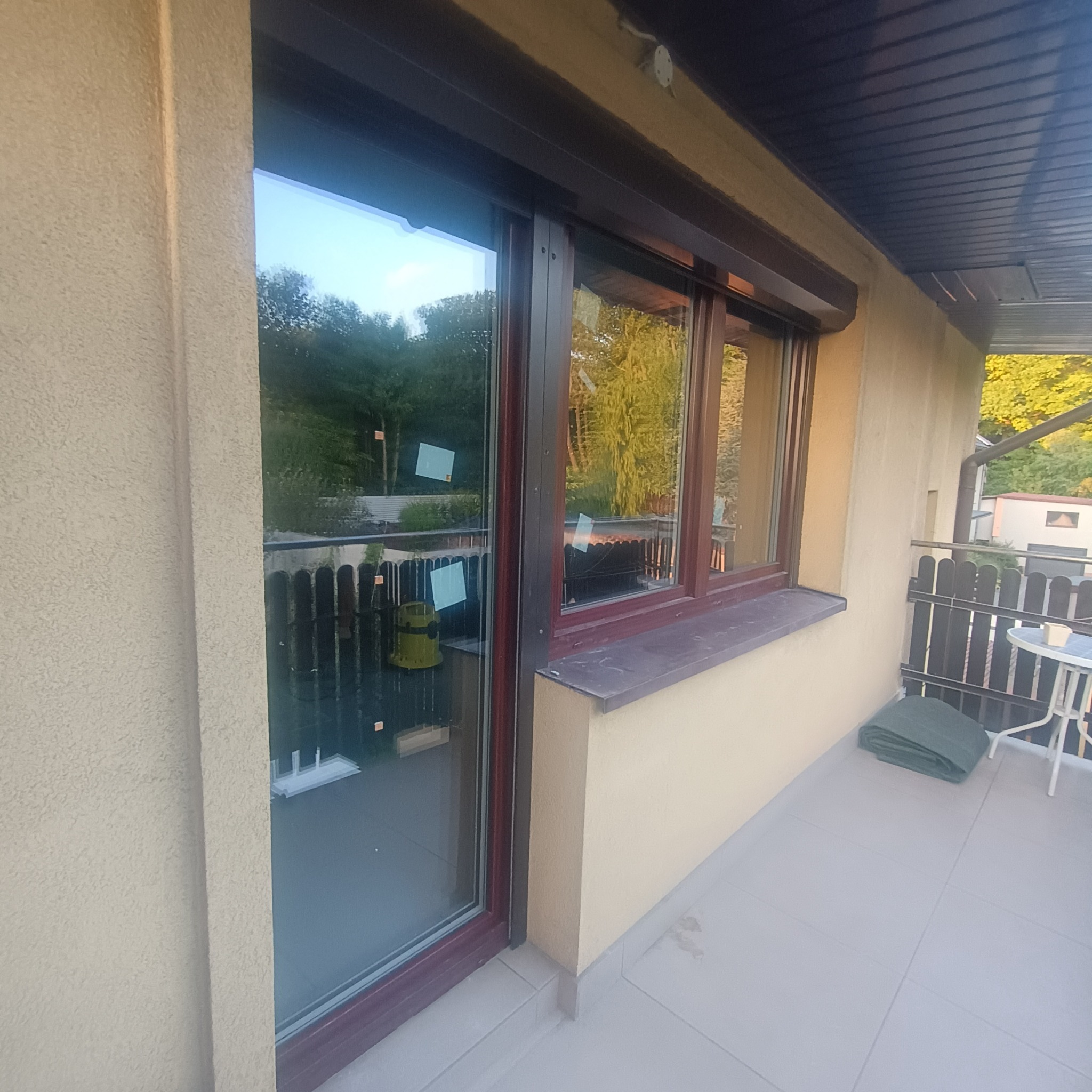 Zewnętrzny widok balkonu z oknem przesuwnym z brązową ramą i zwiniętą roletą. W oknie odbija się zieleń drzew i błękit nieba. Jasna elewacja budynku.